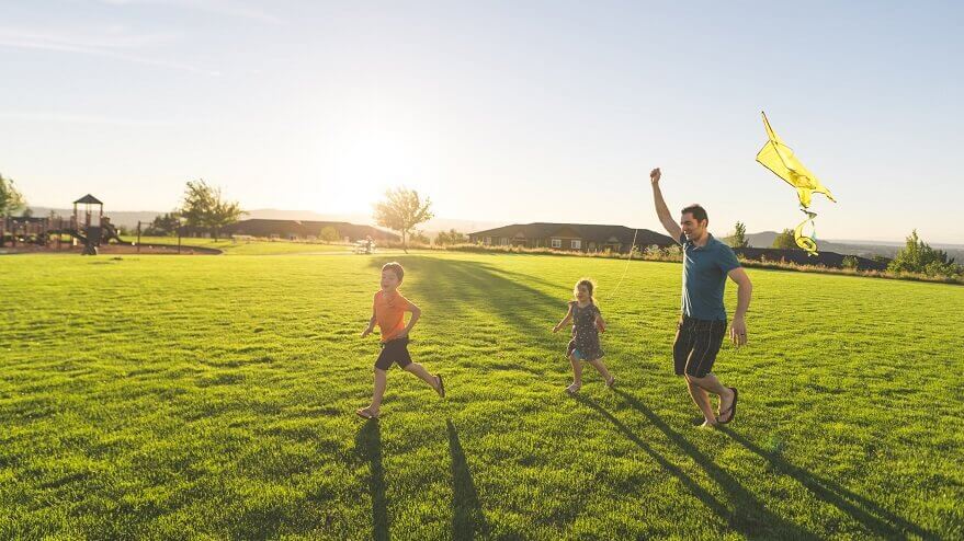 A father and his two young kids fly a kite in a sun-kissed field of gras.